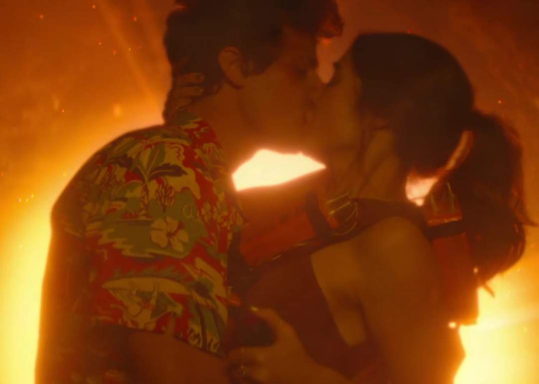 A man and woman kiss passionately in front of a burning ball of color.