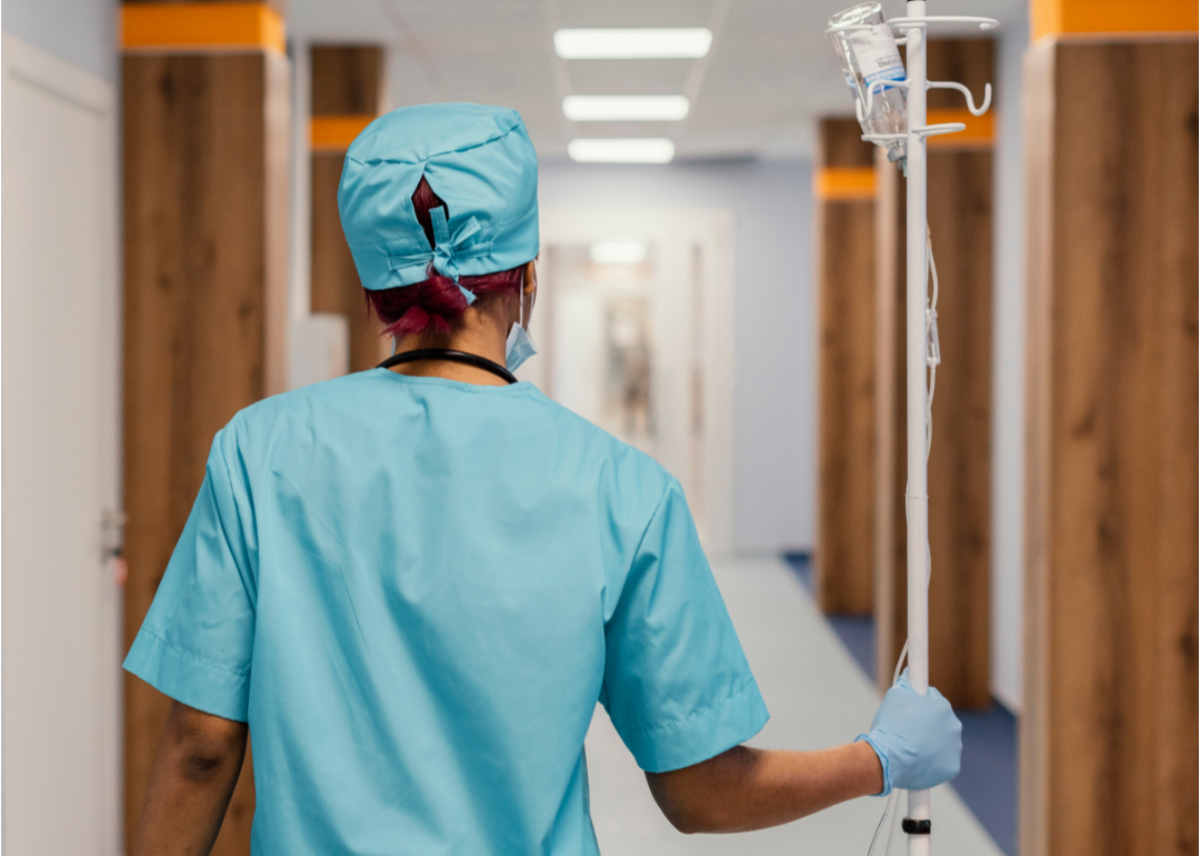 A worker in green scrubs walks down a hospital hallway pushing an IV bag.