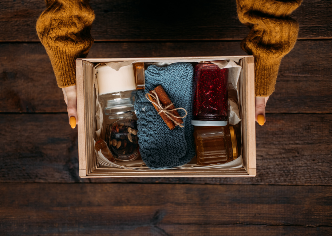 Two hands holding a wooden crate filled with cozy goods including a candle, jar of honey, jar of raspberry preserves, and a blue knitted pair of socks topped with twine-wrapped cinnamon sticks.