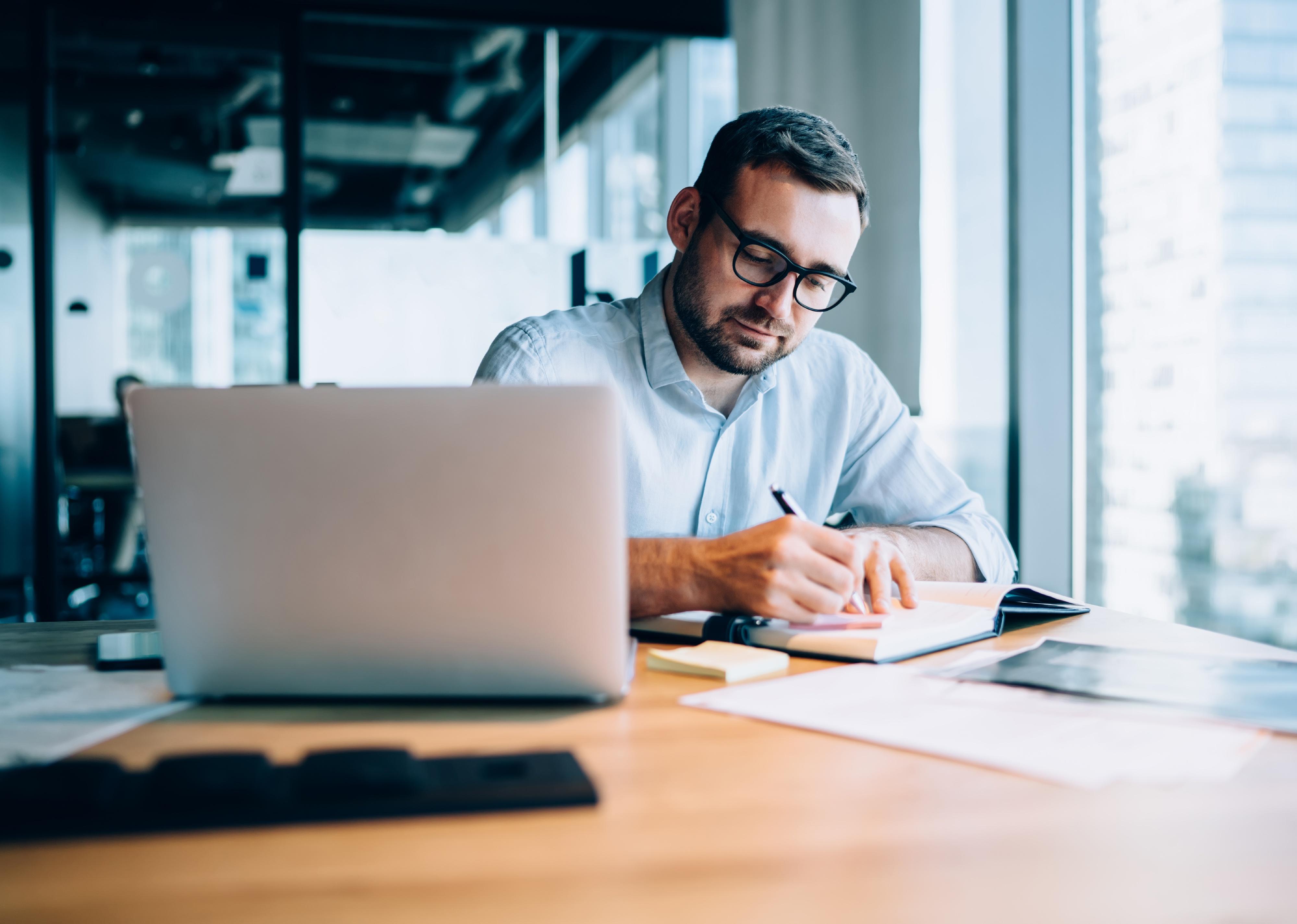 Man writing in book next to laptop