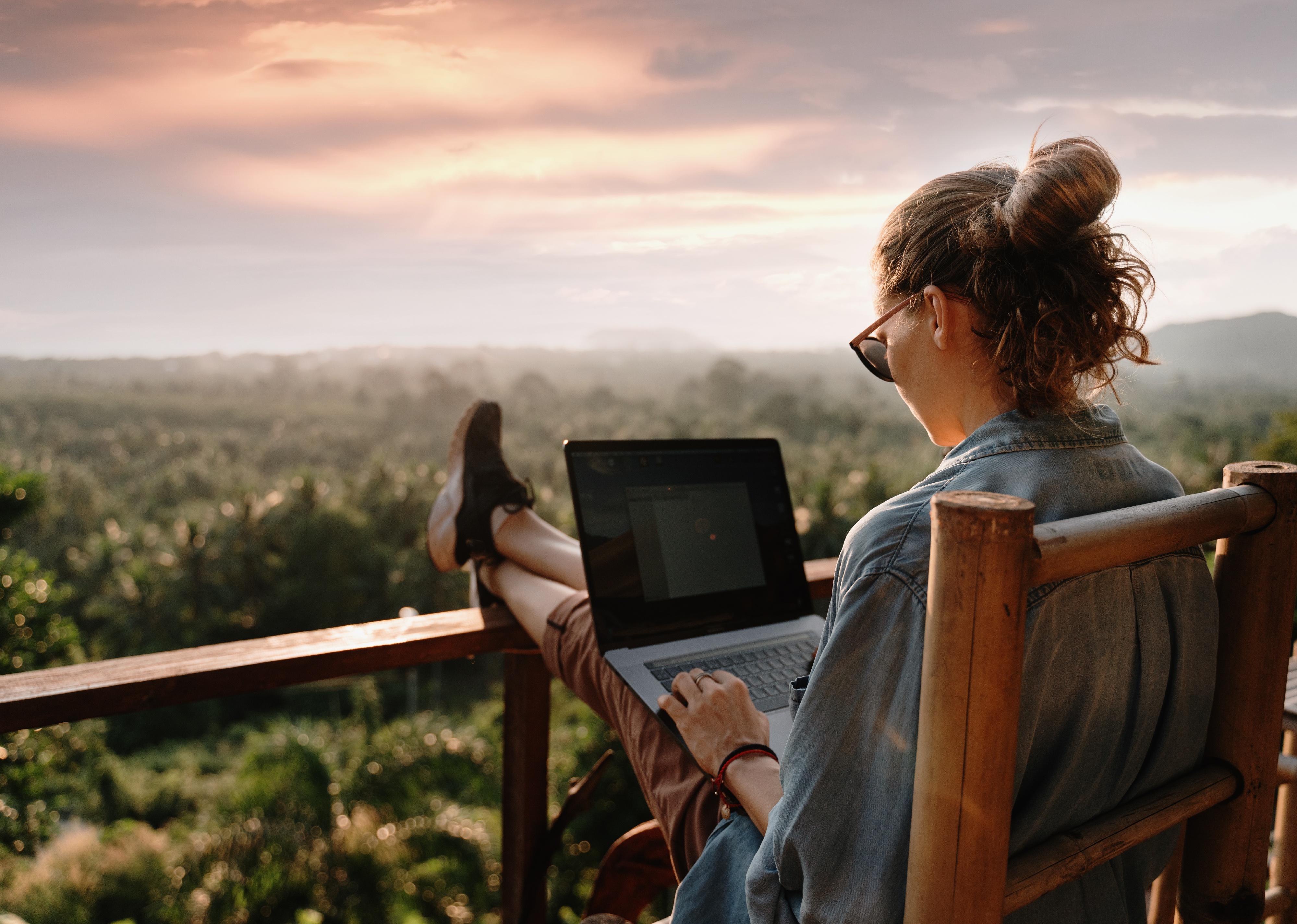 A woman works on her laptop from a balcony overlooking trees