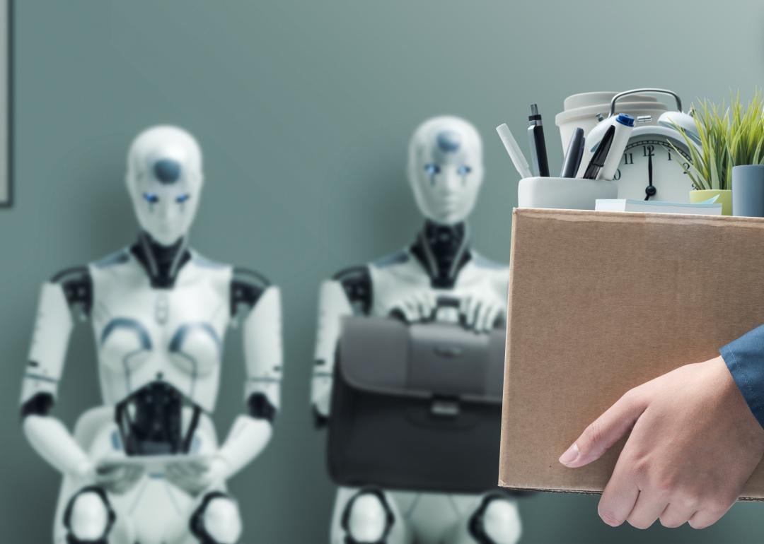 Office worker holding a box with belongings and leaving office, with AI robots waiting for job interview in the background.