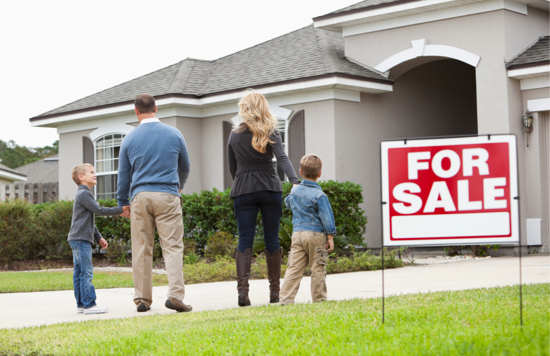 Family looking at house from the exterior with 'for sale' sign in the grass.