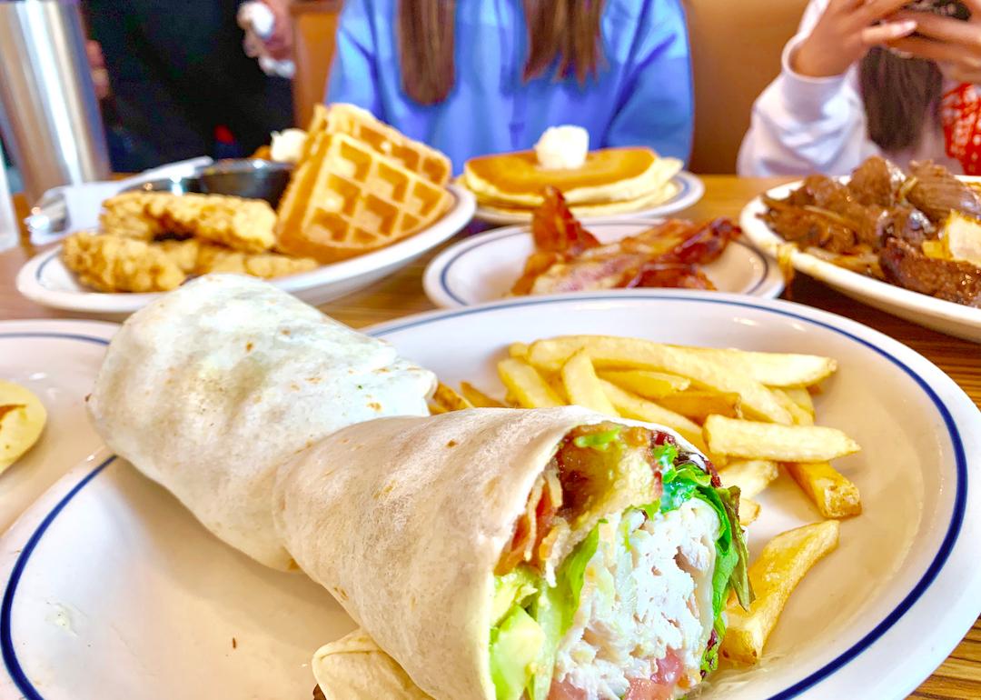 A table at IHOP with plates of brunch items, including a chicken and bacon wrap with fries, chicken and waffles, bacon, and a stack of pancakes.