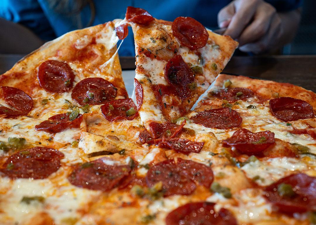 Slice of appetizing pepperoni pizza with melting cheese is being up and picked up from whole pizza by a person off screen.