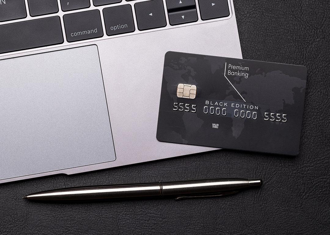Black premium bank card lays on a silver macbook laptop computer next to the trackpad, with a shiny fancy pen on the table.