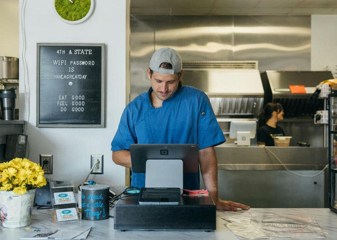 Person wearing a blue shirt entering items into a tablet cash register behind the counter with a kitchen and sign int he background.