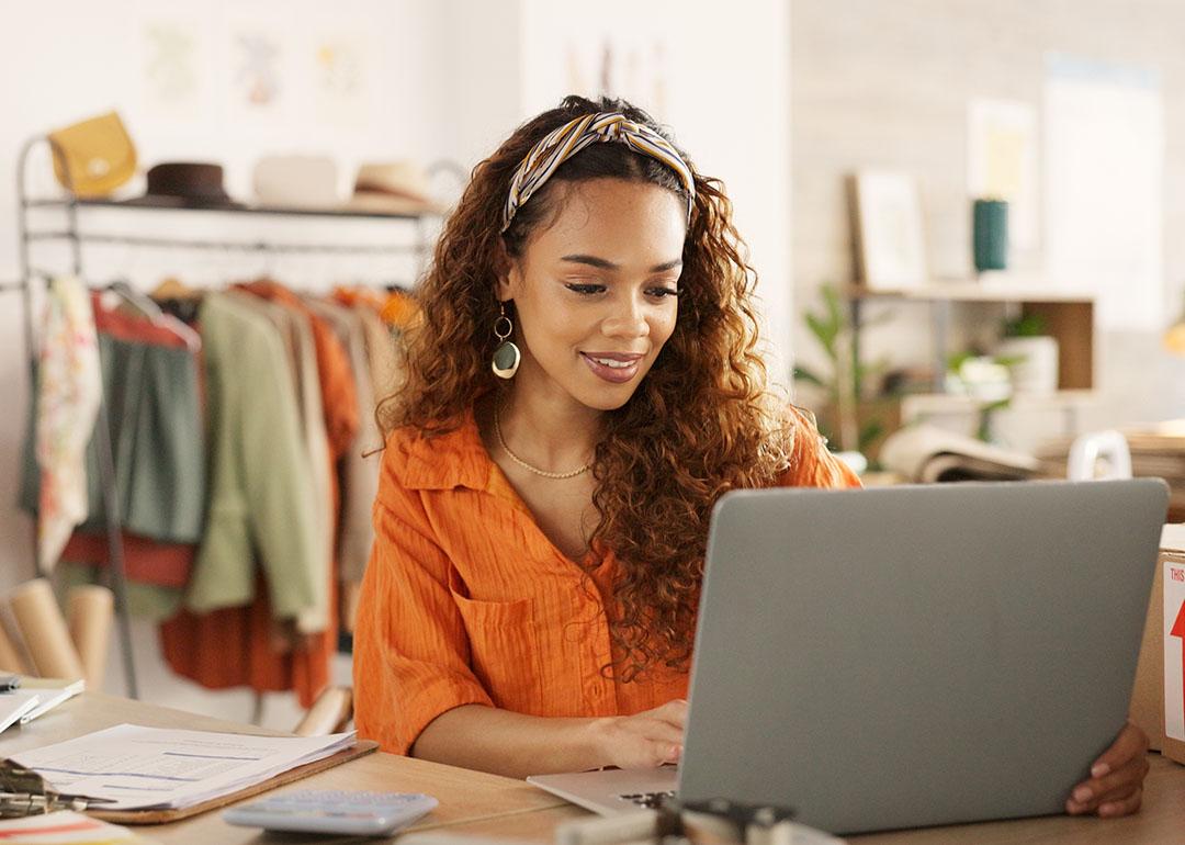 Black woman using laptop in clothing store working on business with rolling rack of clothes behind her.