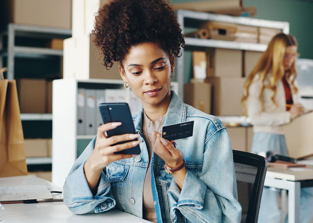 Black woman entrepreneur looking at a business credit card and holding smartphone inside an office with boxes and shelves and a blurred blond person in the background.