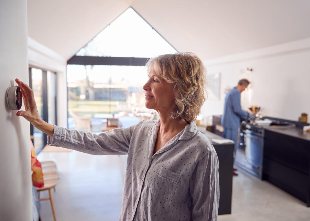 Middle-aged woman wearing pajamas adjusts modern thermostat in a bright house with a vaulted ceiling while a person wearing blue pajamas is seen in the kitchen counter in the background.