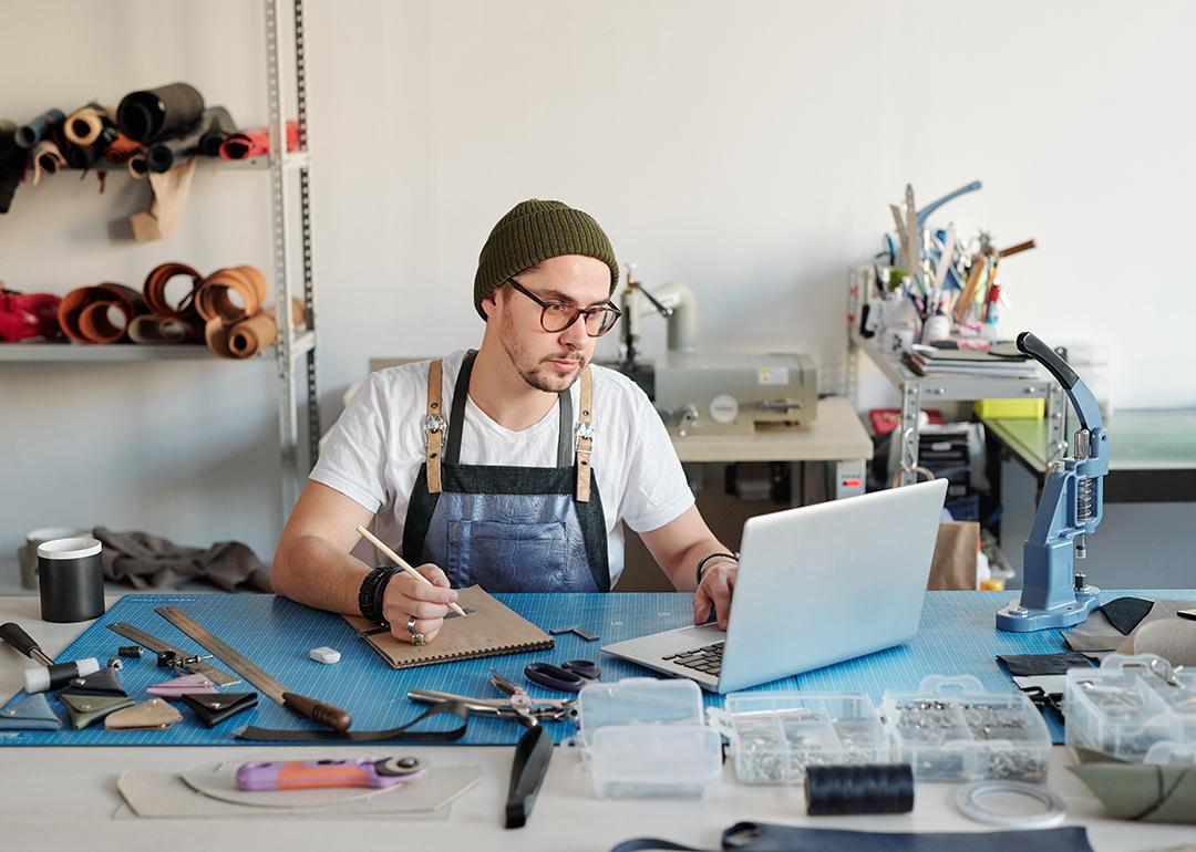 A leather goods craftsman using a laptop and notebook to start his production process.