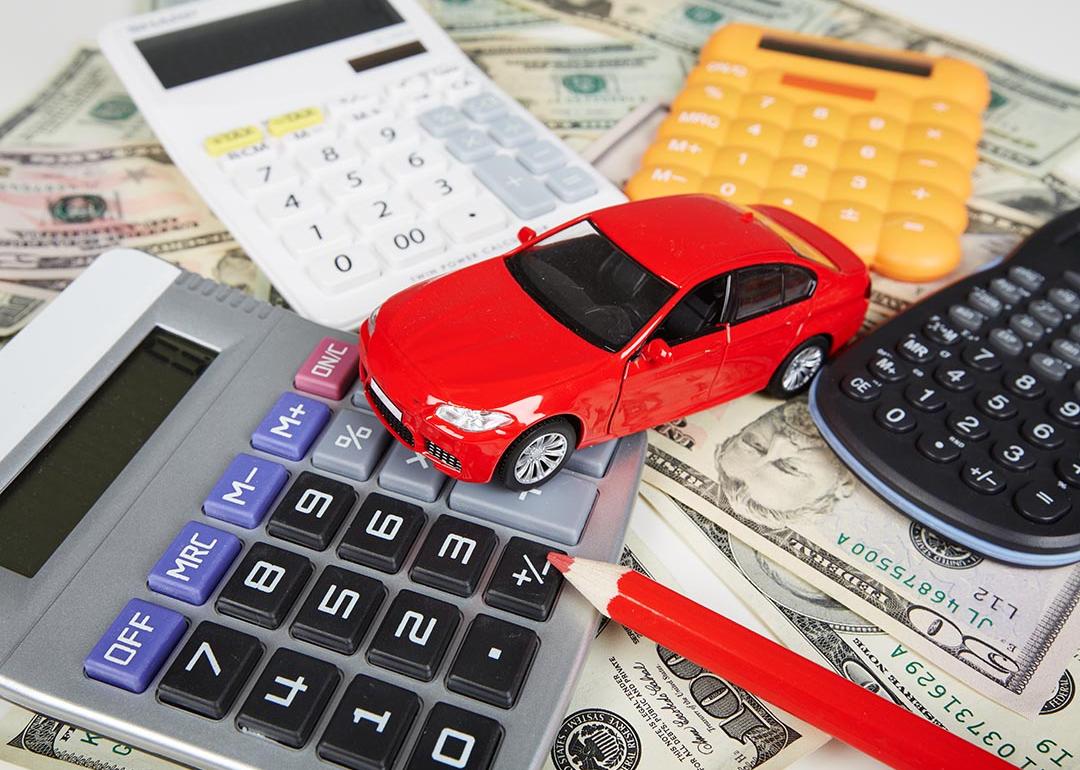 Red toy car surrounded by four calculators and a red pencil sitting on top of U.S. currency to represent the fees required when purchasing a car.