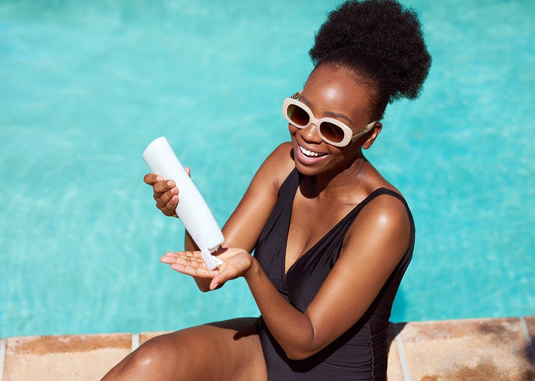 Black woman wearing black suit and white-framed sunglasses applies sunscreen sitting by pool in summer sun