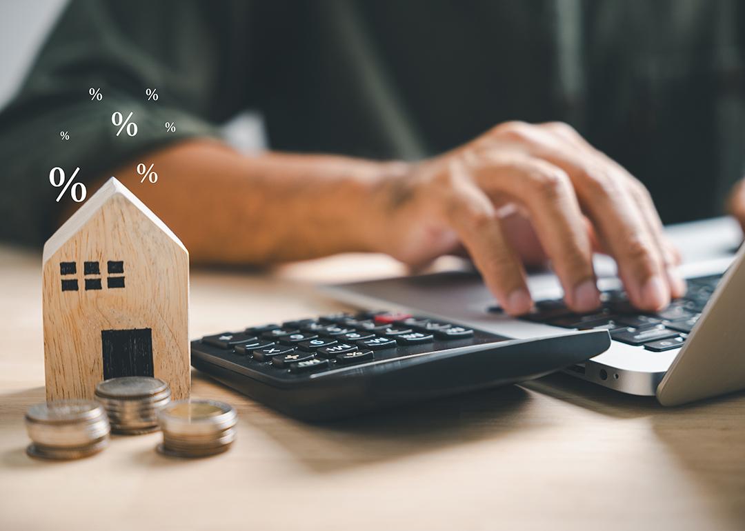 A person using a laptop and calculator with a wooden figure of a house and coins beside him.