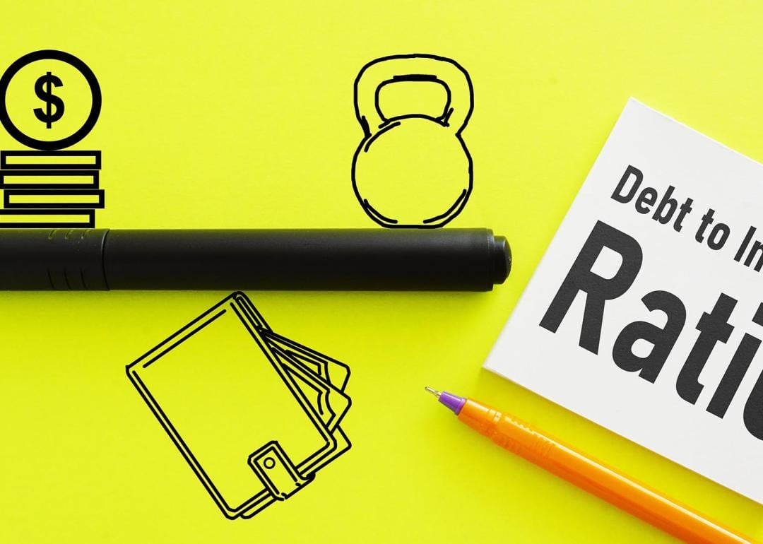 Icons representing money and debt on opposite sides of a line, balancing on a wallet symbolizing income on a flourescent yellow background, with a white post it saying "Debt to Income Ratio."
