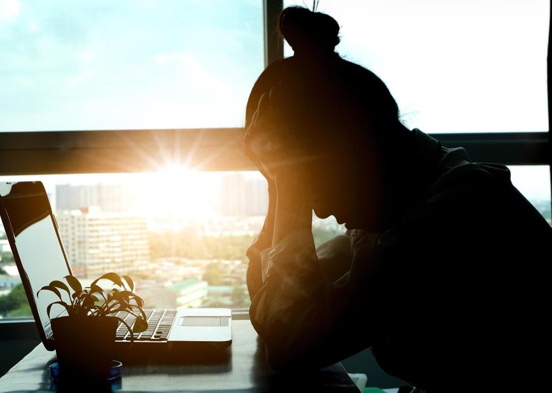 Silhouete of person sitting at desk in front of laptop, with head in hands backlit by sunset shining through office window in background.