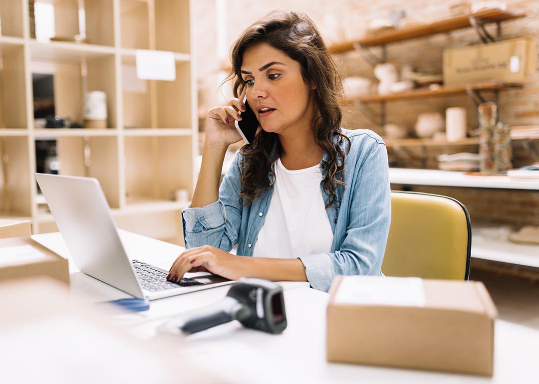 A businesswoman speaking on the phone while using a laptop in her small business production area.