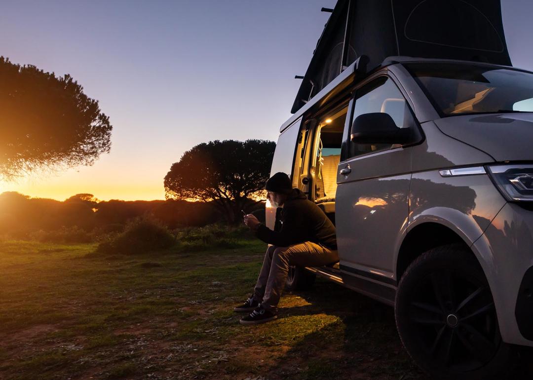 Silhouetted digital nomad sitting in his white transporter camping van, backlit by the setting sun near the California coast.