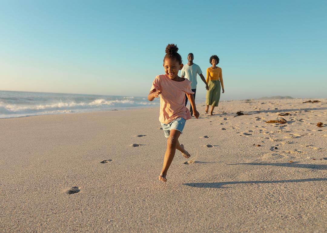 A young girl running on sand against parents walking on a beach.