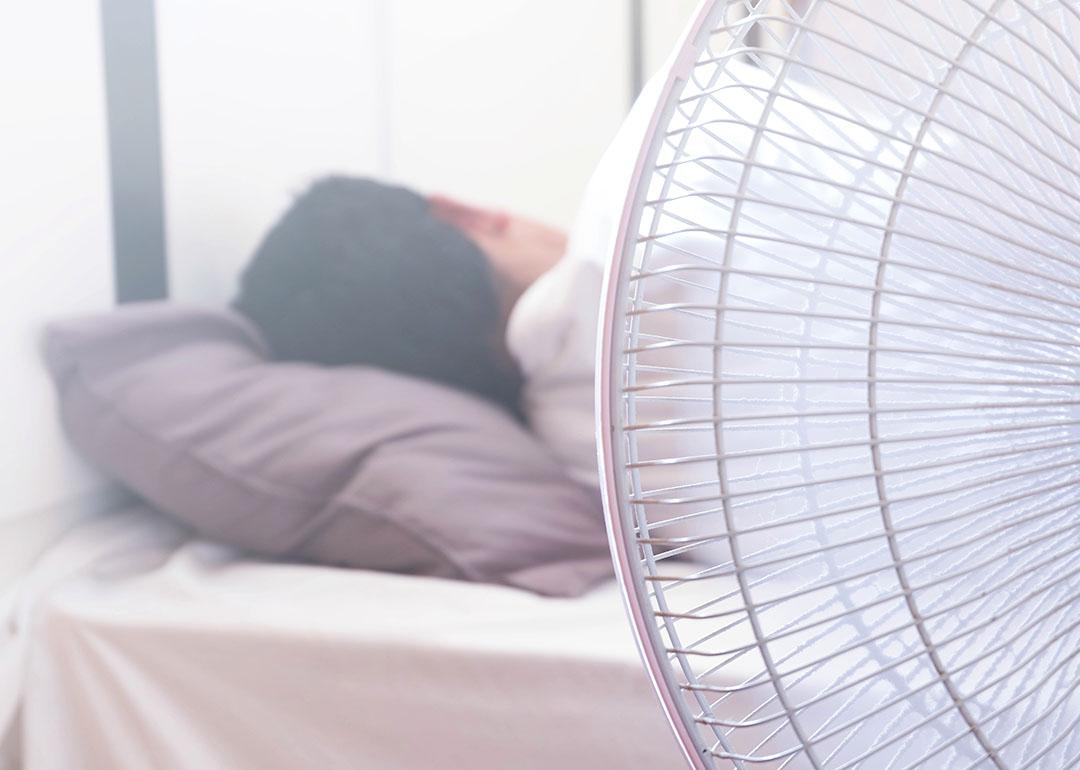 A white electric fan in the foreground in front of a person sleeping in bed, facing away.