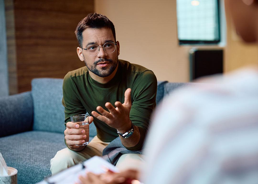 Person wearing long sleeve forest green top and glasses sits on a couch holding a glass of water and gesturing to a person with their back to their camera.