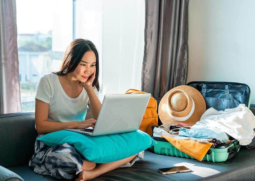 Young woman using a laptop beside an unpacked suitcase preparing for a trip.