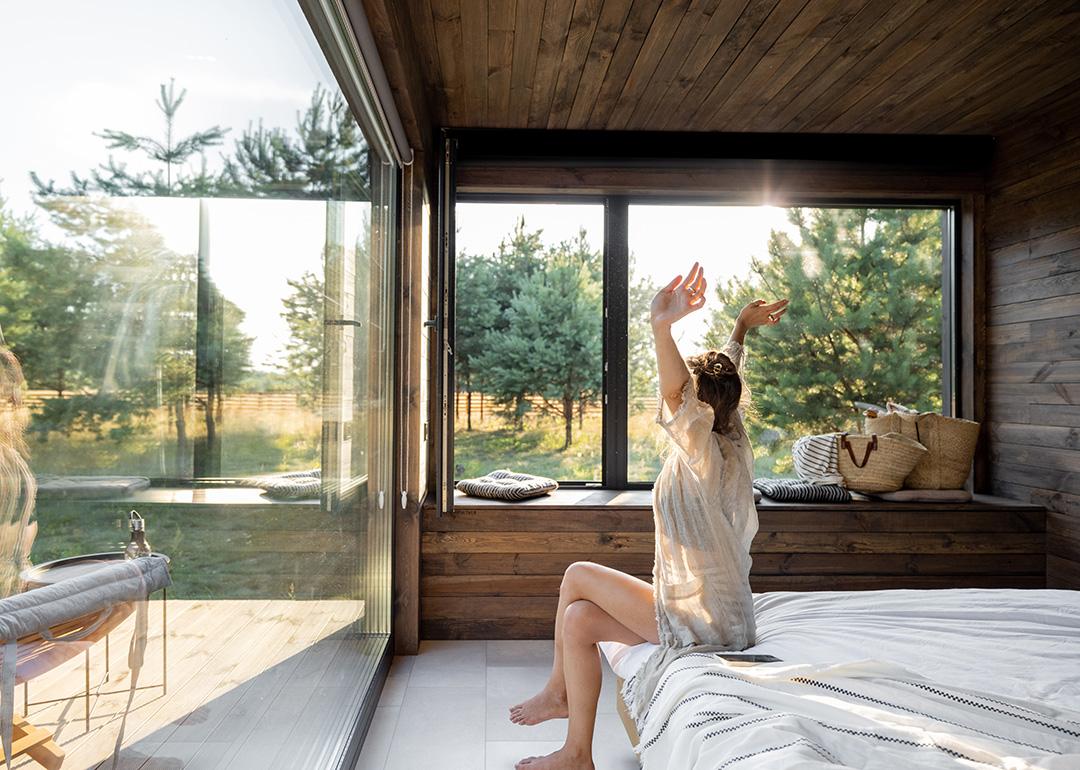 Woman stretching as she is waking up in a modern country house with glass windows.