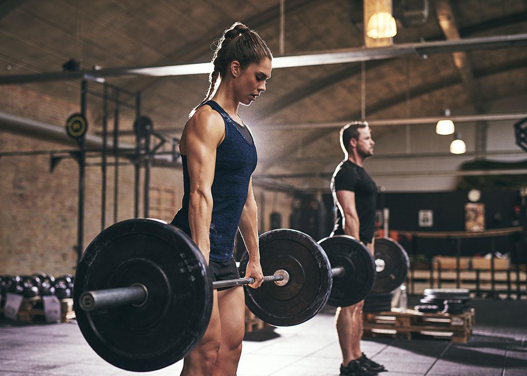 Strong woman and in foreground lifting bar bell with weights with man in background gym.