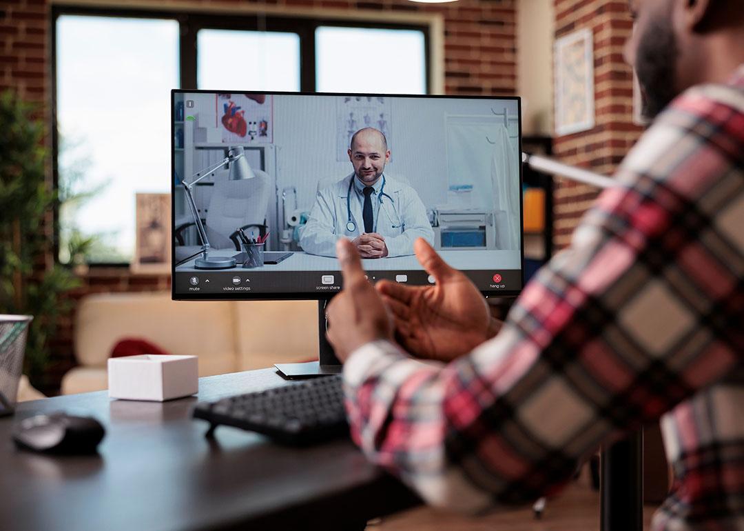 Computer screen showing telehealth call with male balding doctor sitting in office with stethoscope around his neck, while person dressed in plaid long sleeve shirt in foreground converses on video call with his back to the camera.