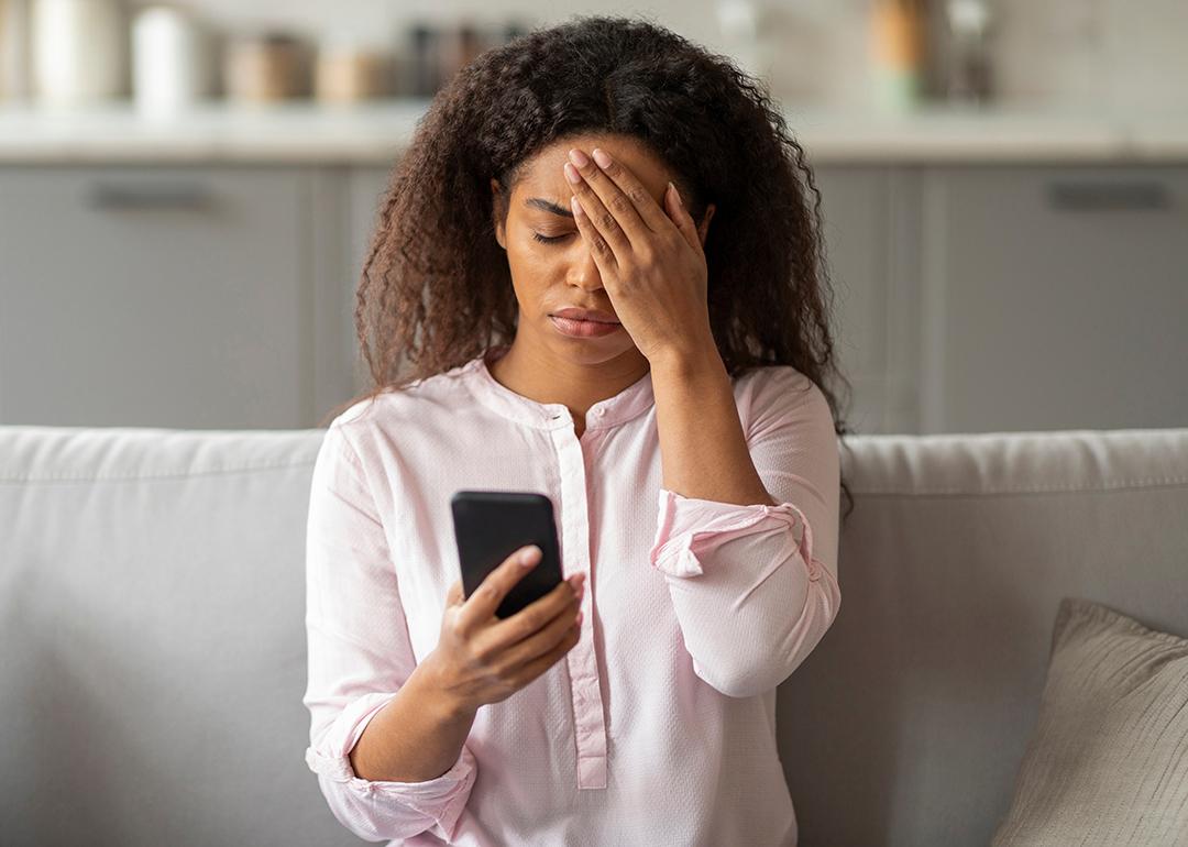 A photo capturing the tension of a young African American woman sitting on her couch, visibly overwhelmed as she looks at her smartphone in a neatly arranged living room.