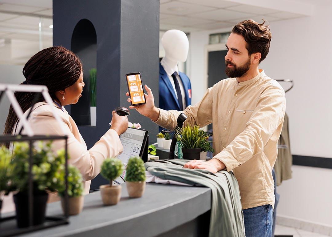 Male customer holding up smartphone with rewards coupon to be scanned by business worker at a retail shop.