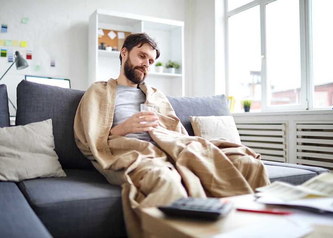 Sick man wrapped into blanket sitting on sofa in front of table with papers while staying at home.