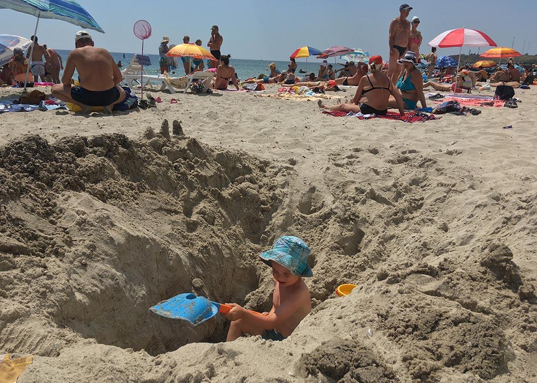 Child wearing a blue hat digging a giant hole in sand at the beach that is higher than him on both sides.