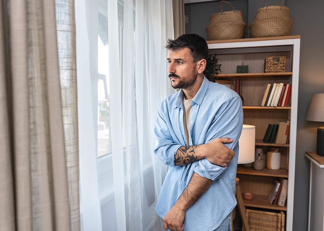 Young sad and depressed anxious looking man wearing a blue button up shirt looking through white curtains out the window.
