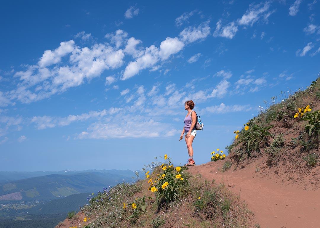 Woman looks out toward view on dirt mountain trail with yellow flowers, hiking in summer with sky and clouds in background near Portland, Oregon.