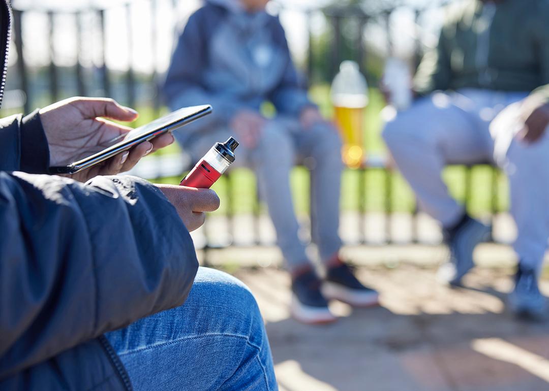 Teenager holding a vape while scrolling on the phone at the park.