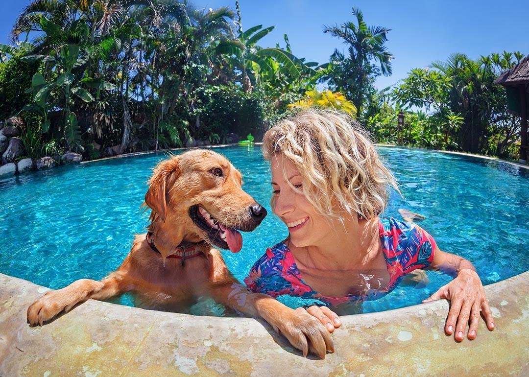 A medium-sized brown dog and blond woman look at each other happily in the pool, with their hands and paws holding onto the edge of the pool.