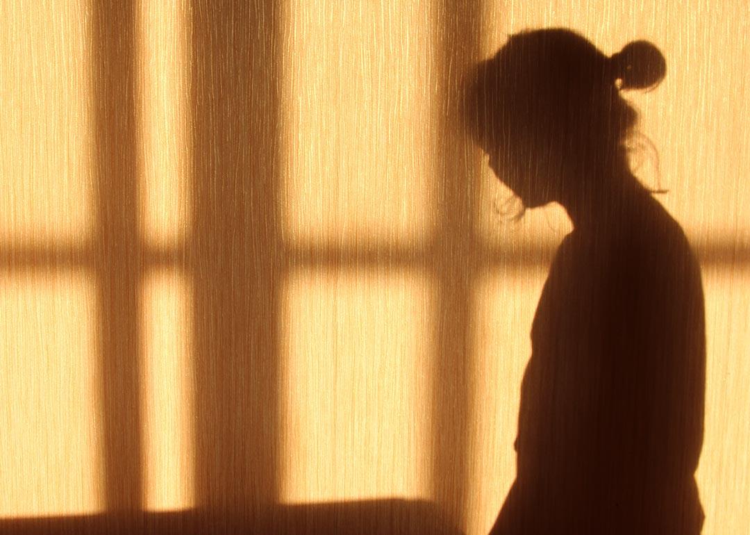 Silhouette of a person with a short ponytail and crossbars of a window on the against a wooden wall. 