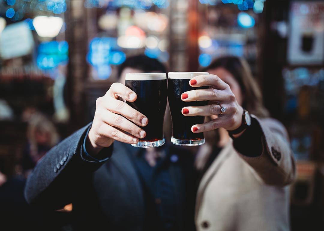Two people holding up a pint each of stout beers to cheers, which obscures their faces from the camera in a bar.