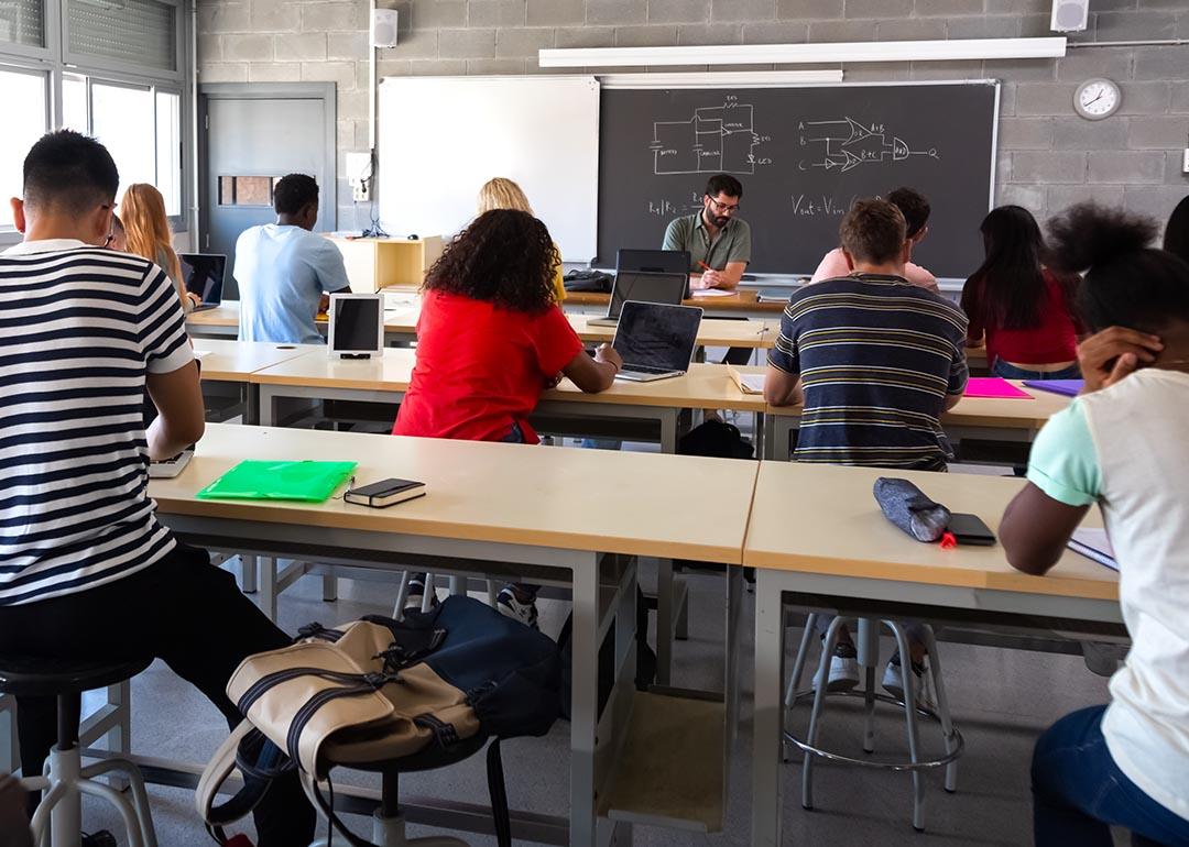 View from back of classroom of multiracial high school students in class using laptops while teacher marks exams.