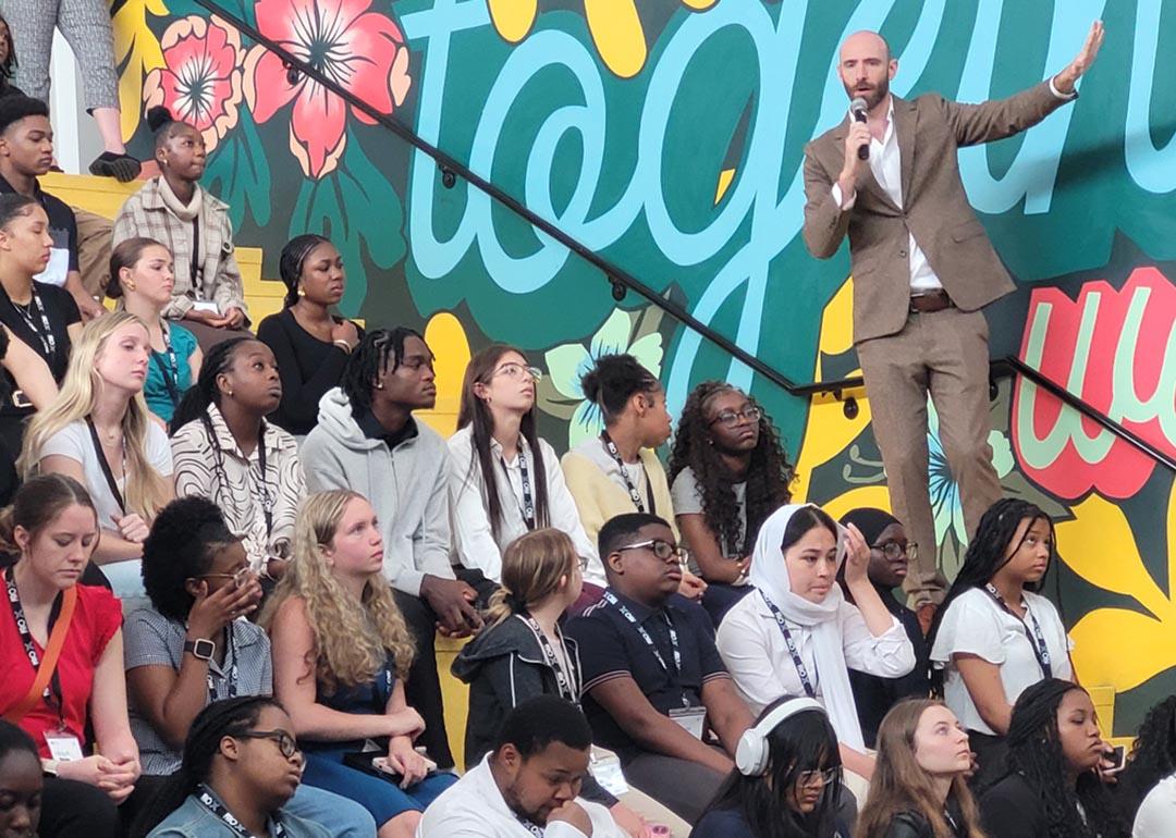 Side view of rows of high school students sitting on bleachers listening to a speaker wearing a tan suit talking into a mic.