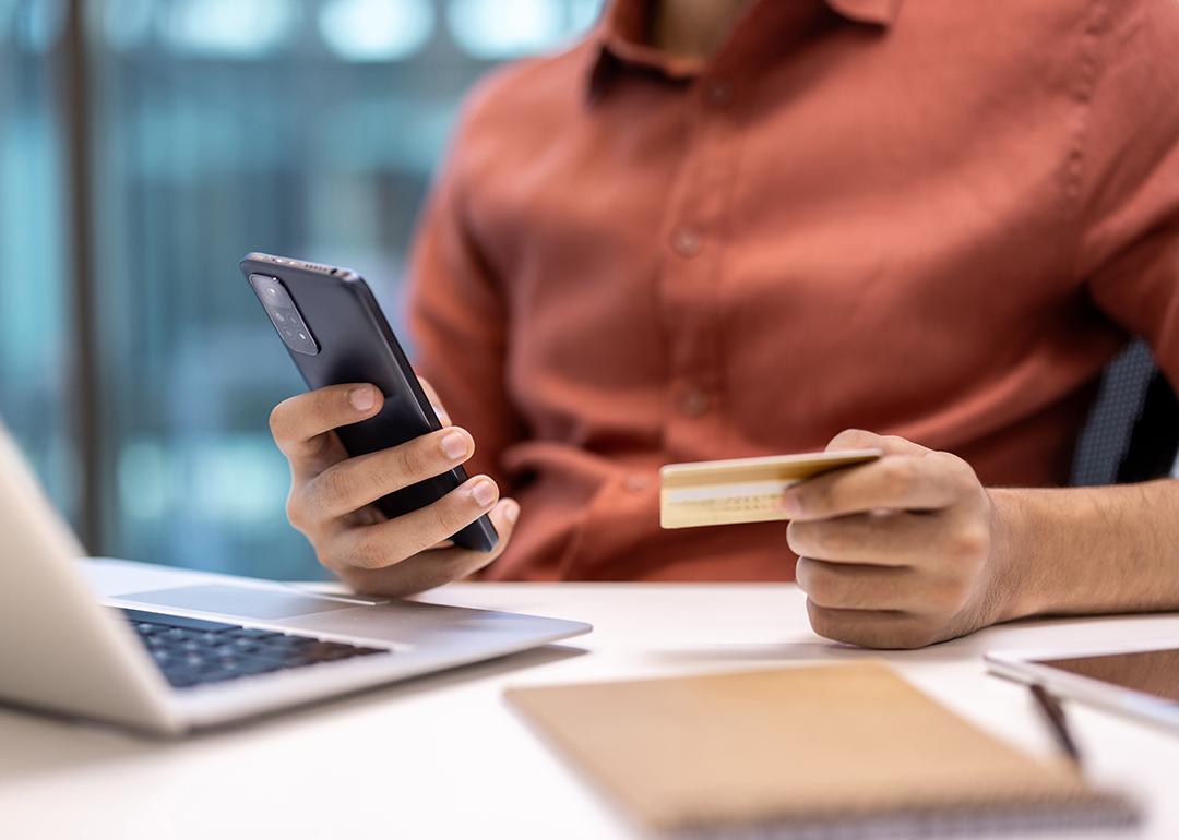 Businessperson holding a credit card while using a phone and laptop at an office desk.