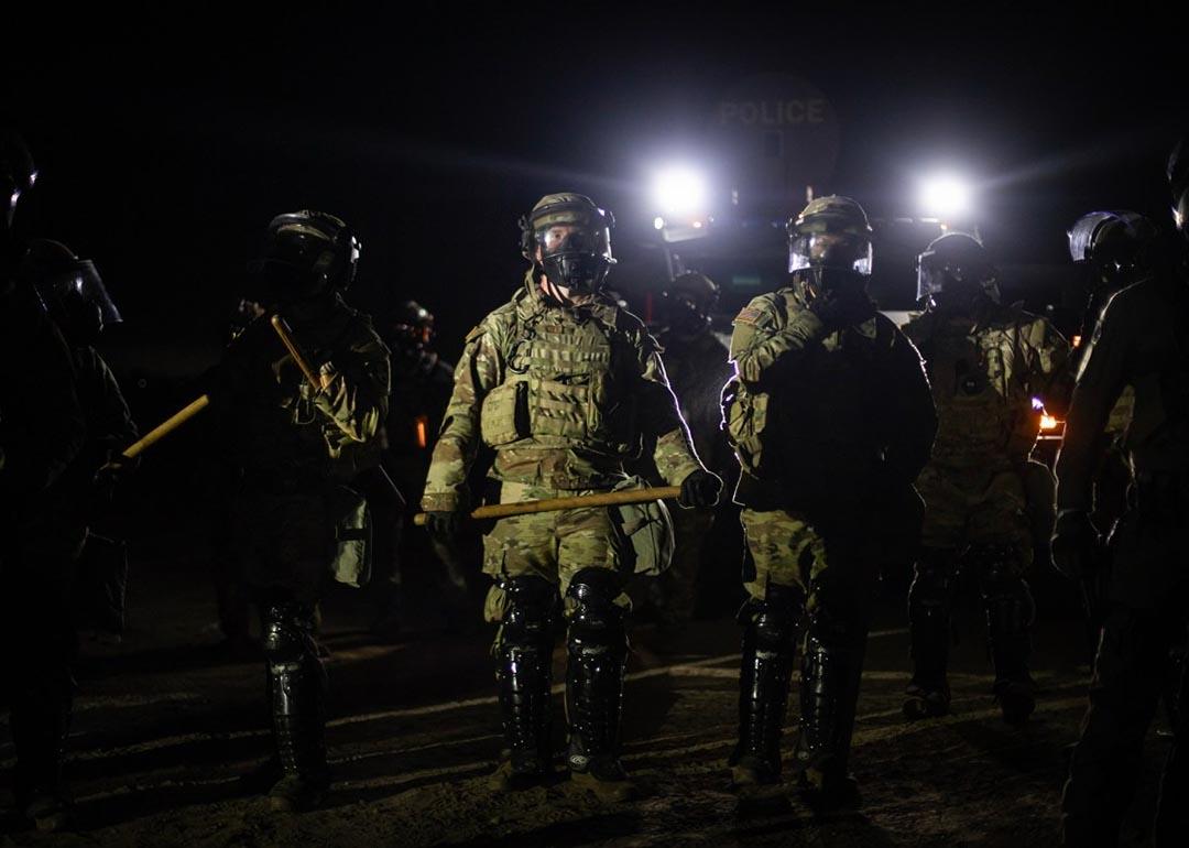 Nighttime view of a line of federal immigration agents in riot gear and face shields facing the camera near Glass House Farms facility in Camarillo, with police spotlights in background.