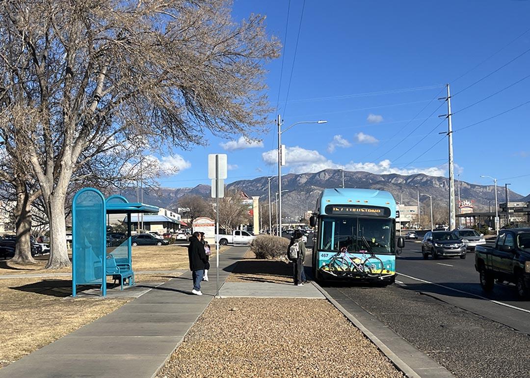 Bus riders board a blue bus in Albuquerque, next to a turquoise metal bus stop and with mountains and cloud in distance.