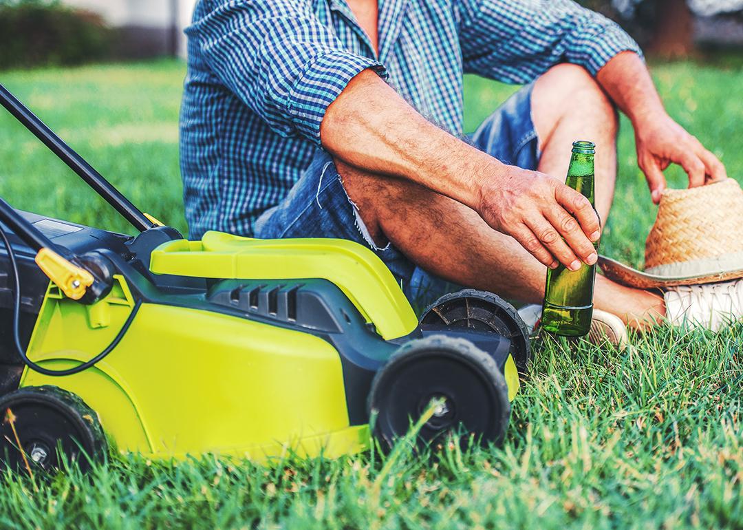 Senior man enjoying a bottle of beer beside a mower in his lawn.
