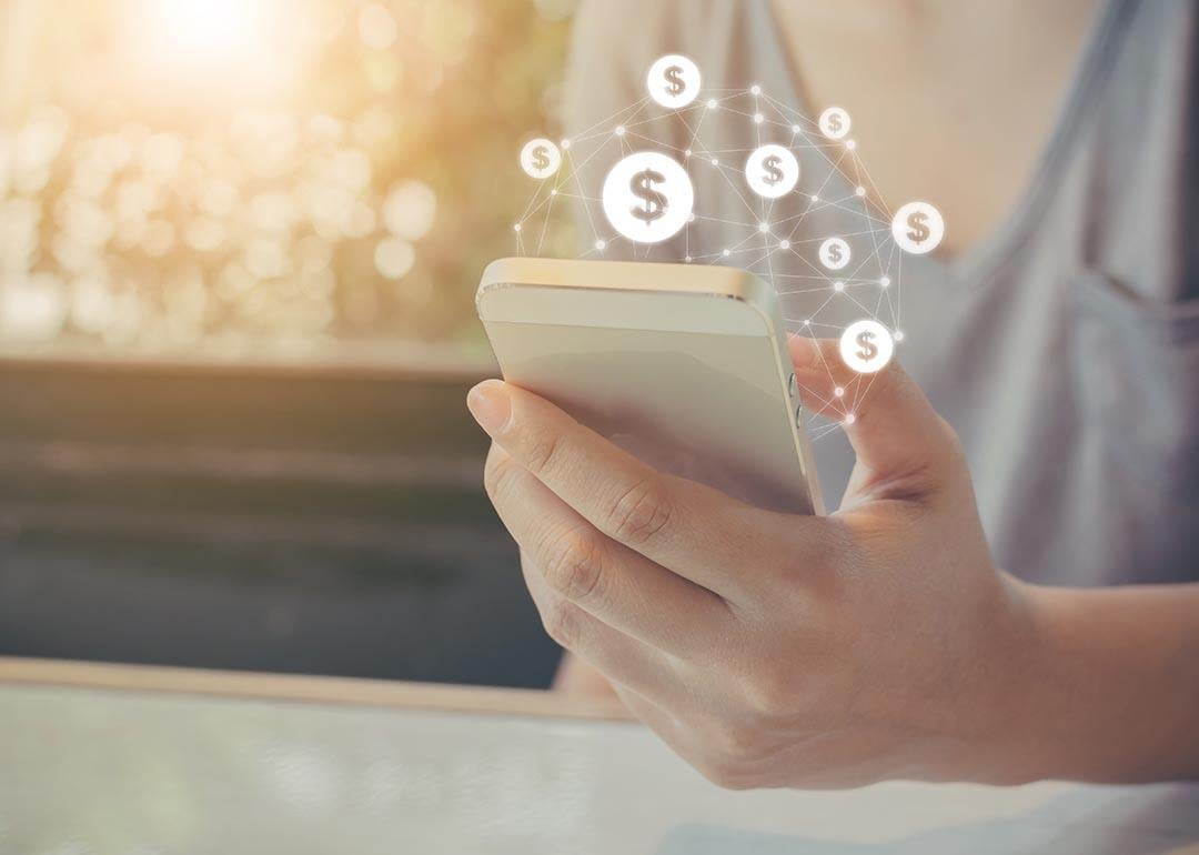 Hand holding mobile phone with dollar sign icons overlaid, symbolizing ecommerce transactions made on smartphones.