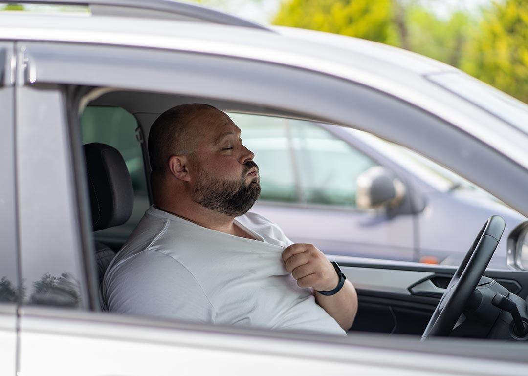 A bald driver with closed eyes seen through the passenger side window pulls at the collar of a white t-shirt and exhales.