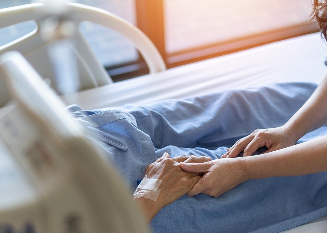 Elderly senior patient in hospital bed, whose face and torse are not visible, has hand held by younger caretaker.