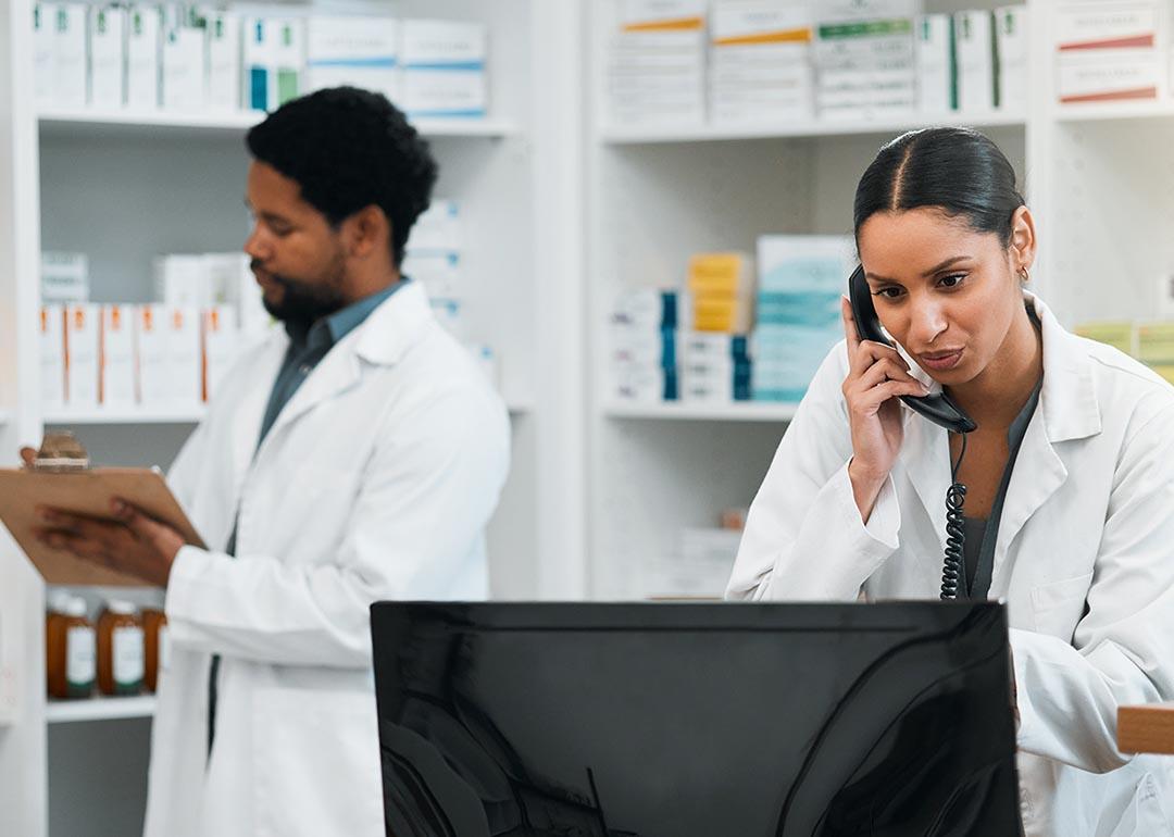 Two pharmacy employees work behind the counter in a pharmacy, one on the phone and looking at a computer and the other writes on a clipboard.