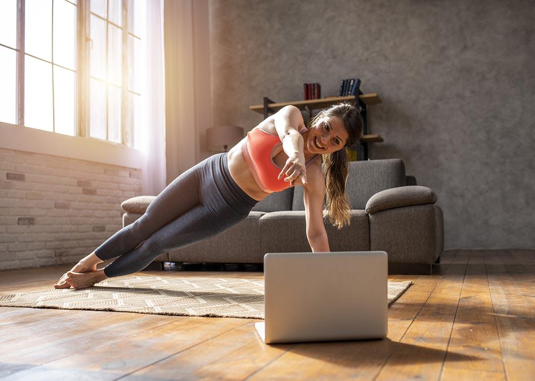 Woman doing a side plank pose on a mat on a wooden floor, following a video on a laptop, with sunlight coming in through large window in the background. 